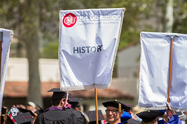 White Banner with the CSU Channel Islands red logo at the top and below the word History in Gray being help up during commencement by someone wearing a black grad cap